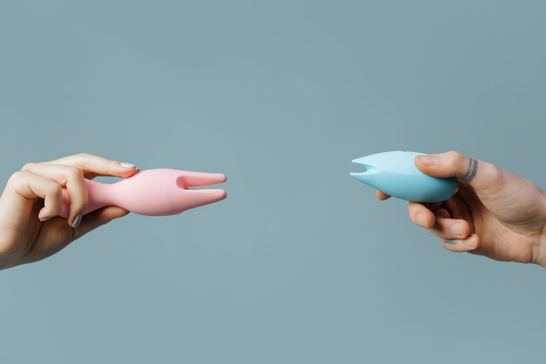 Close-up of hands holding pink and blue silicone clitoral vibrators against a pastel background