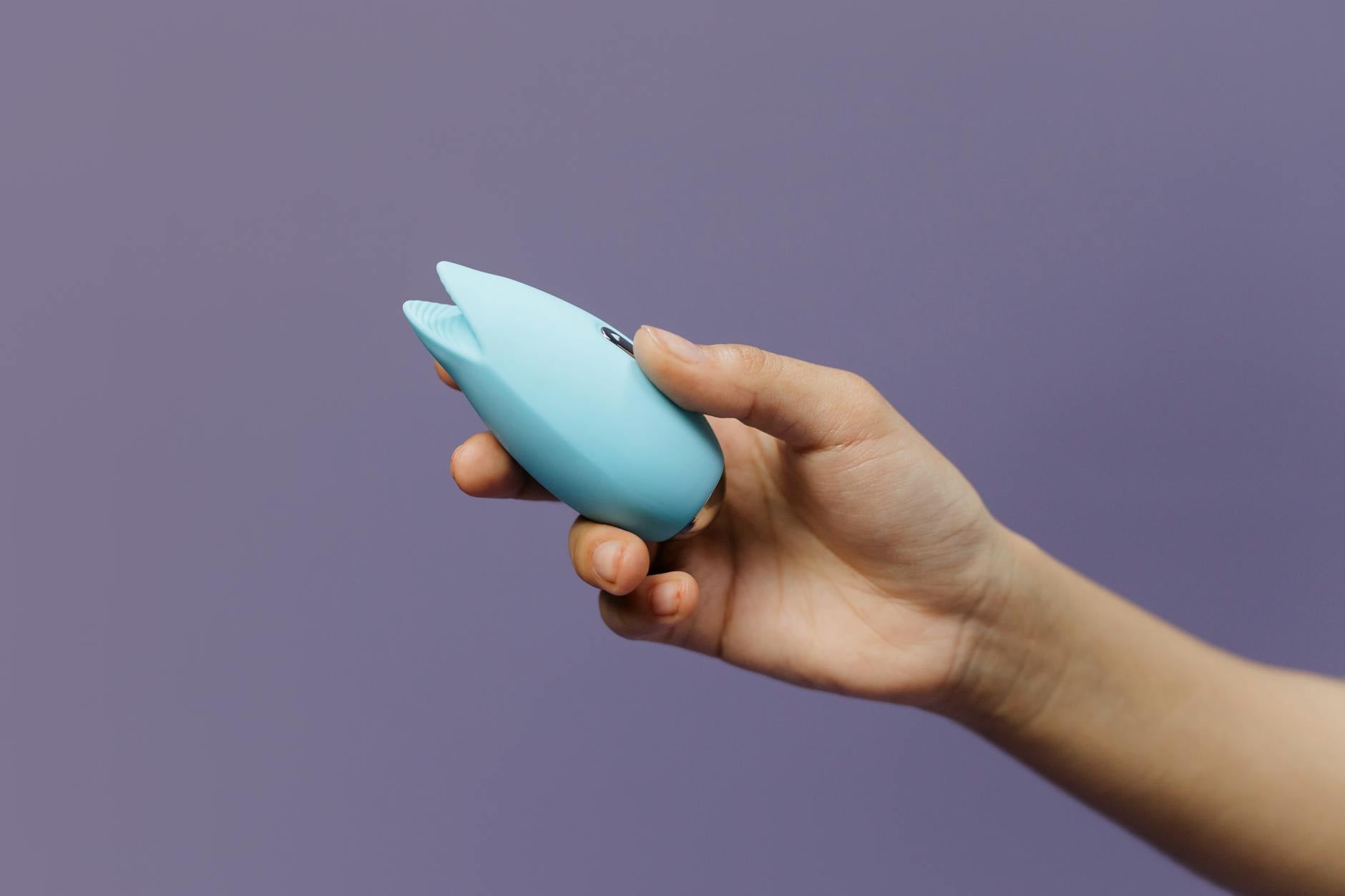 A hand holding a blue silicone clitoral vibrator against a purple background